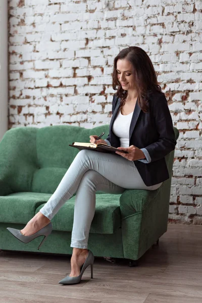 portrait of a business woman sitting on the sofa shelf looking writing in a notebook in a casual suit