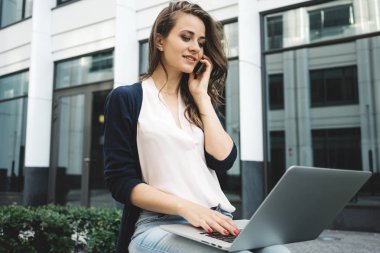 Genç kadın serbest müşteri ile cep telefonuyla konuşmak ve internet bilgi arayın. Kız öğrenci kent yönlendirmek, kullanım modern dizüstü bilgisayar üzerinde açık havada lisansüstü çalışma yapmak