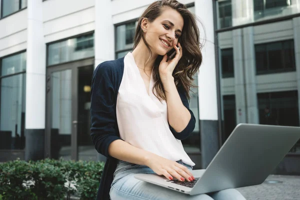 Business woman sits in urban park and typing text on modern laptop and talk by mobile phone. Success female student discussing about graduate work on smartphone