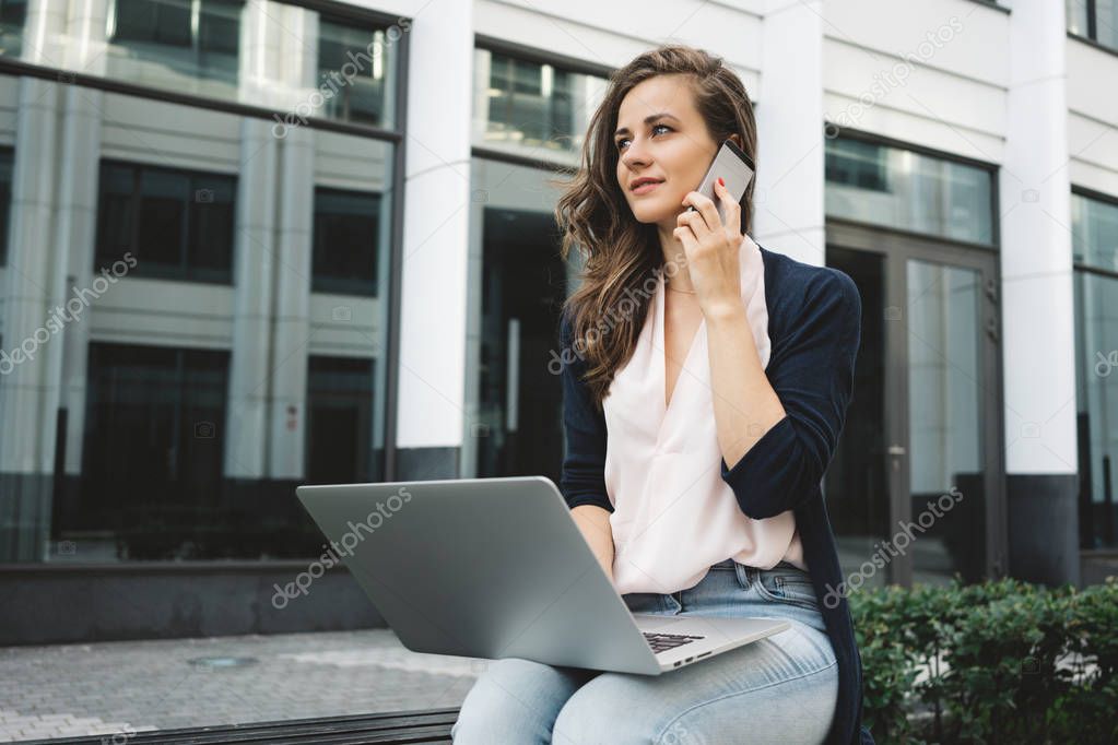 Female freelance working with laptop on street in business district and discusses work by phone with business customer
