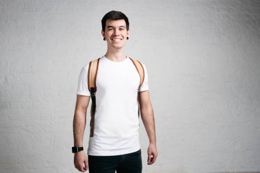 Genç şık gülümseyen adam boş beyaz t-shirt ve sırt çantası. Stüdyo dikey, yatay