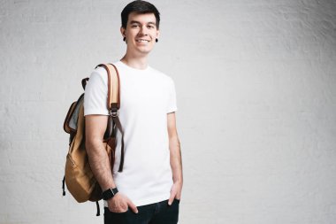 Gülümseyen genç adamın giydiğini boş beyaz t-shirt ve sırt çantası, boş duvar, yatay stüdyo yakın çekim