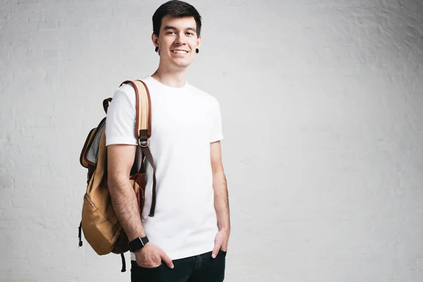 Smiling young man wearing blank white t-shirt and backpack, empty wall, horizontal studio close-up