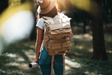 Sevimli bayan şapka, sırt çantası ve konumu harita üzerinde el ormandaki ağaçlar arasında günbatımında arkadan görünüm. Lens flare etkisi