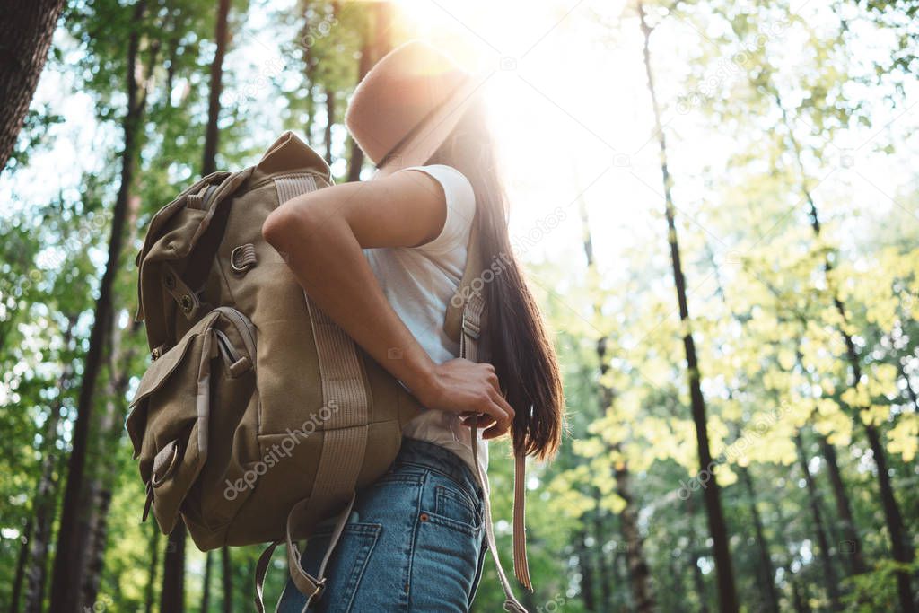 Back view on traveler backpack and hipster girl wearing hat. Young brave woman traveling alone among trees in forest on outdoors. Lens flare effect