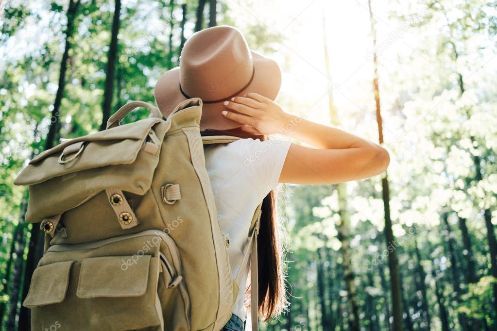 Happy cute traveler woman wearing backpack and hat hiking among forest on outdoors at sunset. Horizontal