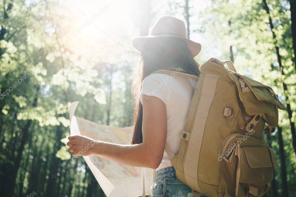 Back view on cute young woman with hat, backpack and location map in hand among trees in forest at sunset. Lens flare effect