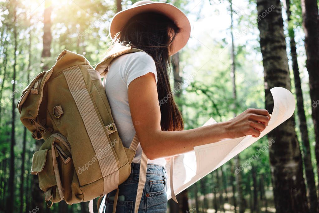 Brave hipster girl traveling alone and looking around in forest on outdoors wearing treveler backpack and hold location map in hand