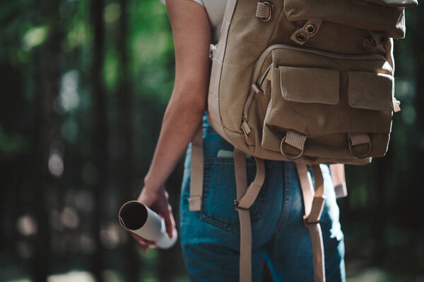 Back view on cute young woman with hat, backpack and location map in hand among trees in forest at sunset. Lens flare effect