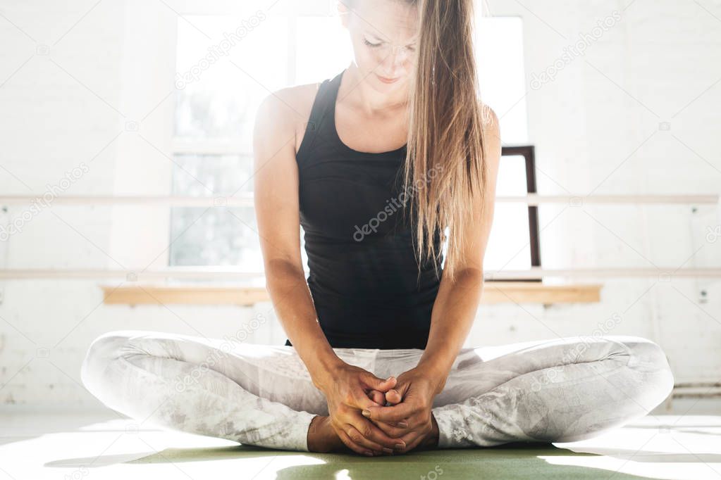 Handsome fit woman doing stretching exercise sitting on yoga mat in sunny white gum. Strong spoty girl warming up before starting workout