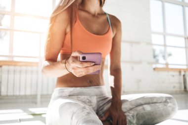 Smartphone cep telefonu ile el sportif kız güneşli beyaz spor salonunda eşofman giyiyor görünümü kapatın. Yoga mat üzerinde oturan kadın uygun ve temizliğini telefonu el sayar kalori fitness uygulama sonrası eğitim