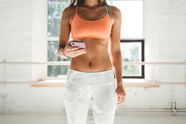 Tanned sporty woman wearing sportwear in white sunny gym hold smartphone in hand and counts calories in fitness application fater workout. Sport and gadget concept