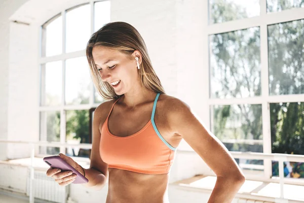 Healthy happy woman with modern headphones hold smartphone in hand. Fitness girl training in white gym and counts calories in application on mobile phone