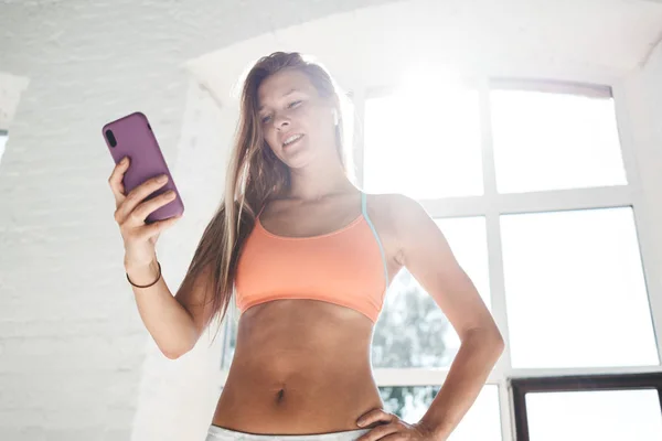 Tanned sporty woman wearing sportwear and wireless headphones in white sunny gym hold smartphone in hand and counts calories in fitness application fater workout. Sport and gadget concept