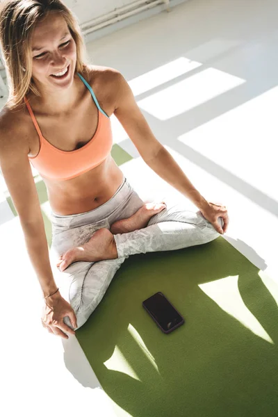 Healthy happy woman with modern headphones hold smartphone in hand. Fitness girl training in white gym and counts calories in application on mobile phone