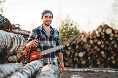 Güçlü el arka planını kereste fabrikası ve ambar ağaçlarının ekose gömlek tutun chainsaw giyiyor oduncu sakallı