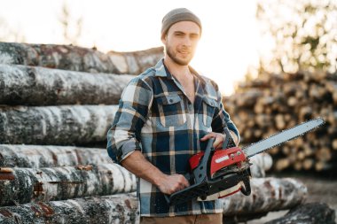 Güçlü el kereste fabrikası arka plan üzerinde ekose gömlek tutun chainsaw giyiyor oduncu sakallı 
