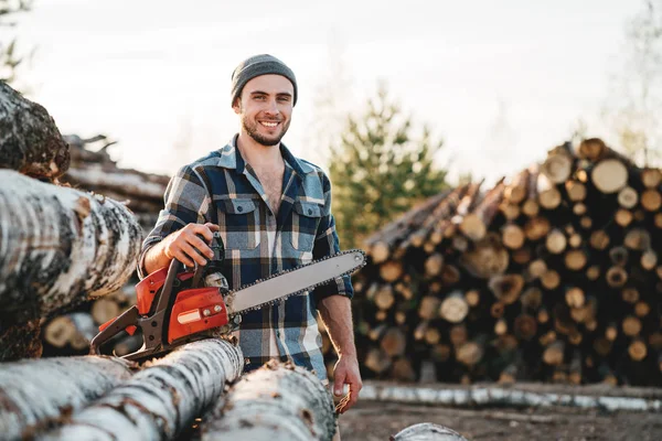 身穿格子衬衫的强胡子樵夫手持电锯在锯木厂和树木仓库的背景下 — 图库照片