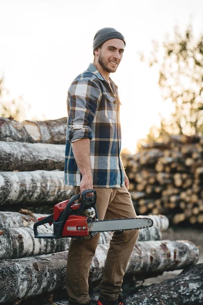 Bearded strong lumberjack wearing plaid shirt hold in hand chainsaw for work on sawmill. Vertical