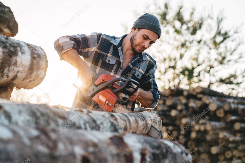 Professional bearded strong lumberman wearing plaid shirt sawing tree with chainsaw for work on sawmill