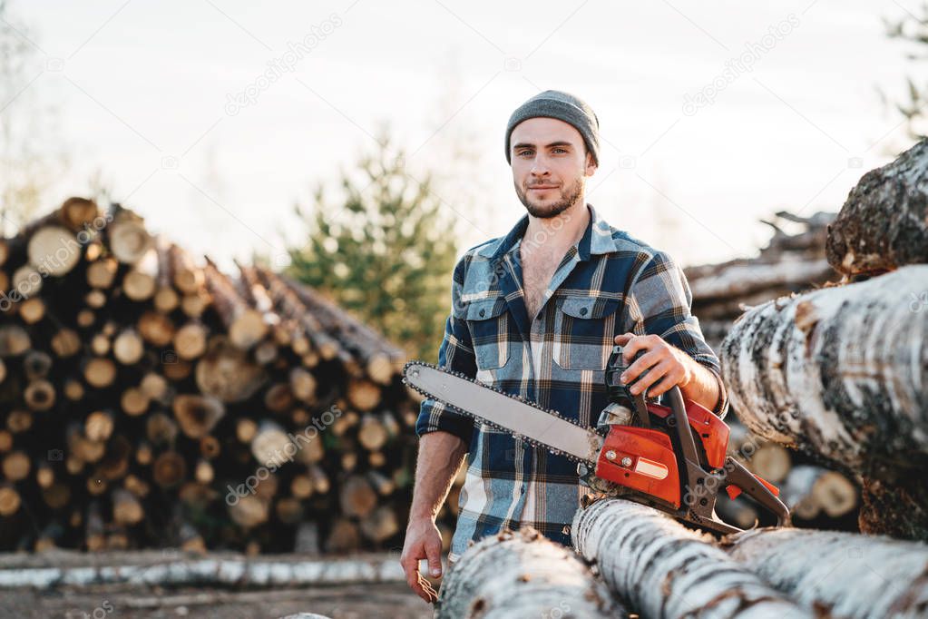 Bearded strong lumberjack wearing plaid shirt hold in hand chainsaw for work on sawmill
