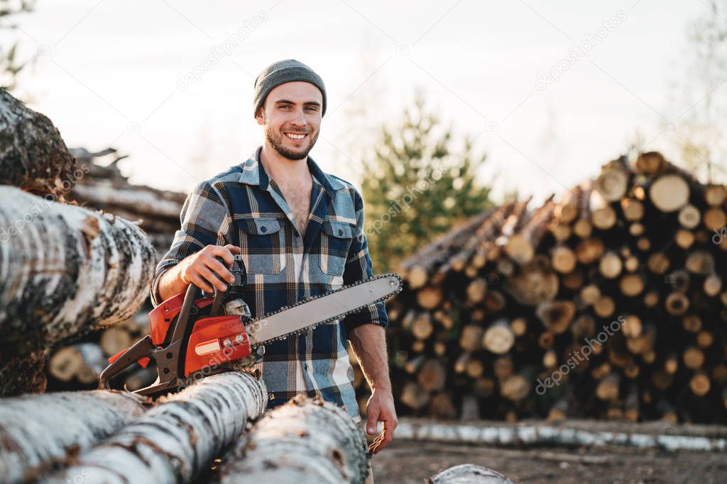 Strong bearded lumberjack wearing plaid shirt hold chainsaw in hand on background of sawmill and warehouse of trees