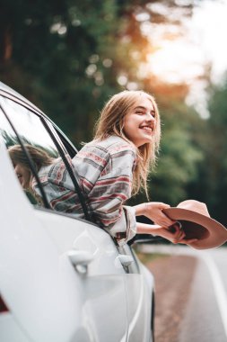Hipster kadın vahşi orman yolda araba ile seyahat. Kareli gömlek ve şapka giyen yakışıklı mutlu kız bir araba direksiyon başında oturan ve penceresinden bakıyor. Yolculuk tutkusu