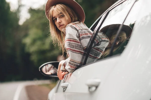 Güzel ve arabada oturan ve penceresinden vahşi yolda seyir kadın seyahat tarzı. Boho kız, yolculuk tutkusu. Kareli gömlek giymiş