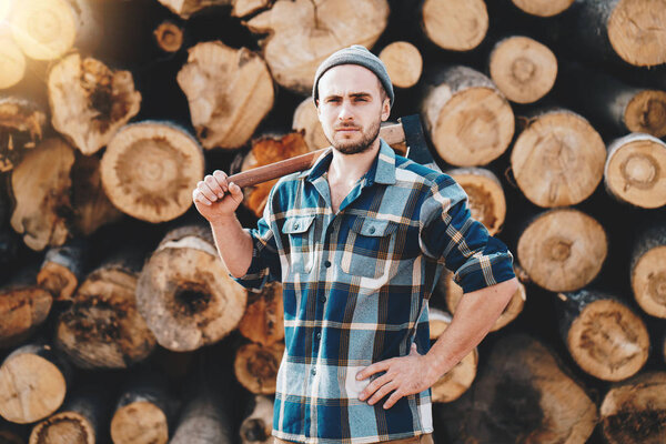 Strong bearded lumberman wearing plaid shirt holds axe on his shoulder on background of sawmill