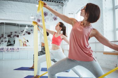 Portre güçlü uygun kadın Eğitim Yoga fitness mat ile birlikte bir grup insan arka plan üzerinde genişletici bant ile poz veriyor. Sağlık ve yaşam tarzı