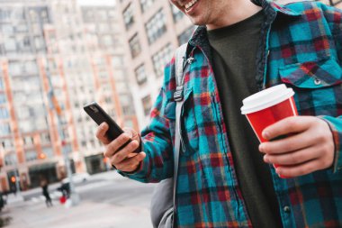 Fincan sıcak kahve ile şehir sokak, cep telefonuna mesaj üzerinde yürüyen adam gülümsüyor. Hippi kentsel görünümü için arkadaşları smartphone cep telefonu ile mesaj yazarak