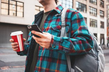 Büyük şehir fincan sıcak kahve ile sokak yürürken ve arkadaşları ile video bağlantı için smartphone kullanarak trendy adam. Casual adam cep telefonu ekranda mesaj yazarak