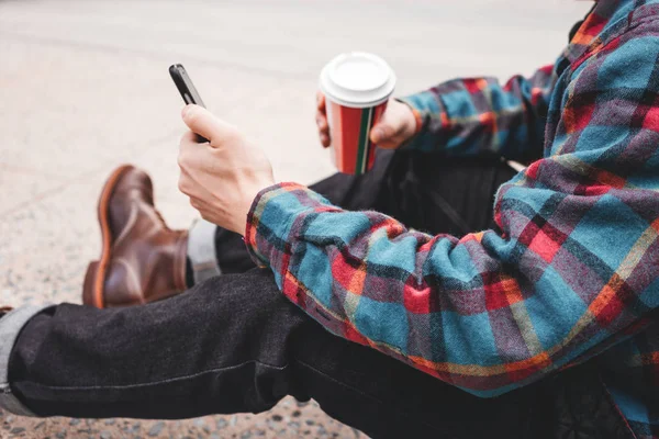 Man in casual clothing hold in hand cup of hot coffee and using smartphone. Young hipster sitting on the Manhattan street and typing text on mobile phone