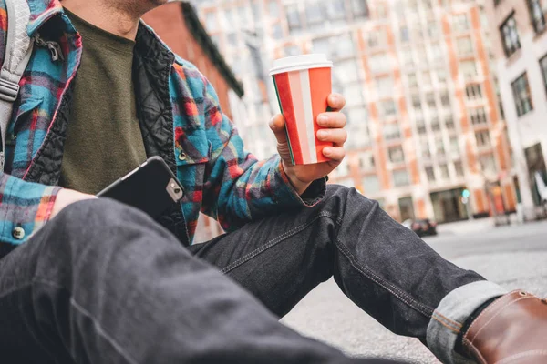 Hipster man sitting on street sidewalk testing with cup of hot coffee and using mobile phone