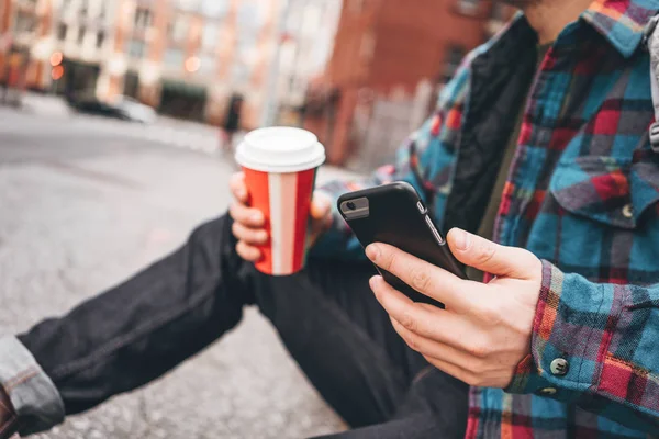 Hipster man resting with cup hot coffee on city sidewalk and texting massage on mobile phone screen. Man using smartphone and sitting on urban sidewalk road