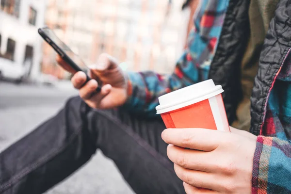 Hipster man sitting on street sidewalk testing with cup of hot coffee and using mobile phone