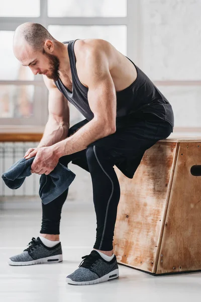 Tired strong man sitting on box at gym. Perspiring fit sportsman resting after hard crossfit workout in light hall. Vertical