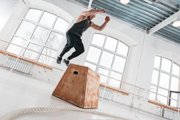 Fit male athlete doing box jump exercise in light hall. Sport man wearing in sportswear is performing box jumps at gym