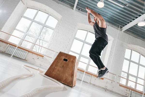 Dynamic shot of fitness male athlete jumping at square box in crossfit gym. Strong man doing jump on wooden squat box at light hall