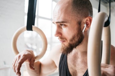 Spor salonunda jimnastik halkaları tutan ve uzağa arıyorsunuz uygun erkek portresi. Fitness adam kablosuz kulaklık yoğun egzersiz yaptıktan sonra yorgun arıyorsunuz
