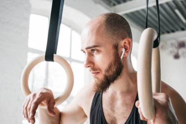 Güçlü kablosuz kulaklık crosstraining yüzük crossfit Salonu'nda holding ile atlet sakallı. Jimnastik yüzük hafif spor salonunda tutarak ve uzağa arıyorsunuz kaslı adam