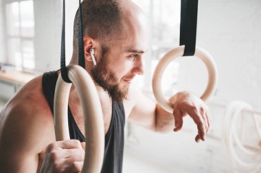 Portre erkek atlet spor salonunda kablosuz kulaklıklar giyen sakallı. Jimnastikçi halkaları tutan ve uzağa bakarak gülümseyen uygun adam