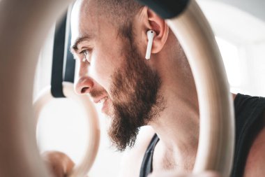 Sakallı erkek atlet spor salonunda kablosuz kulaklıklar giyen jimnastik halkaları aracılığıyla görünümü kapatın. Jimnastikçi halkaları tutan ve uzağa bakarak gülümseyen uygun adam