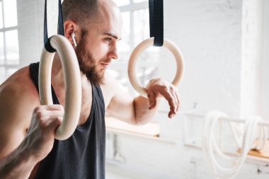 Jimnastikçi halkaları tutan ve uzağa bakarak yorgun uygun bir adam. Sakallı atlet spor salonunda egzersiz Kablosuz Kulaklık takmış 