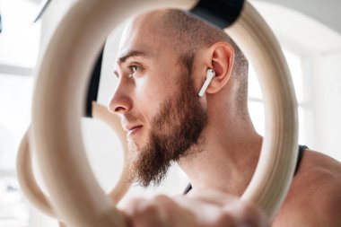 Sakallı erkek atlet halkaları aracılığıyla dinlenme ve hafif spor salonunda jimnastik halkaları tutan kablosuz kulaklık ile görüntüleyin. Adam rahatlatıcı yoğun egzersiz yaptıktan sonra uygun