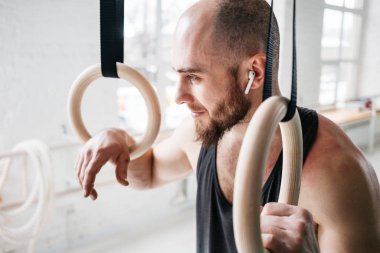 Sakallı erkek atlet dinlenme ve jimnastik yüzük hafif spor salonunda holding modern kulaklıklarla. Uygun adam rahatlatıcı jimnastik kez çaldıktan sonra yoğun çalışma üzerinde