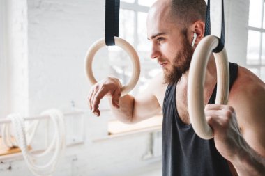 Dinlenme ve hafif spor salonunda jimnastik halkaları tutan kablosuz kulaklık ile erkek sakallı atlet. Uygun adam rahatlatıcı jimnastik kez çaldıktan sonra yoğun çalışma üzerinde