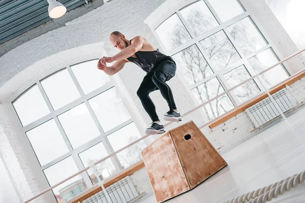 Fit sportsman doing jump exercises on wooden box in cross workout gym. Strong male athlete jumping at box in workout hall