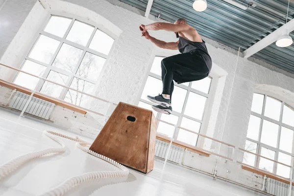 Fit sportsman doing jump exercises on wooden box in cross workout gym. Strong male athlete jumping at box in workout hall