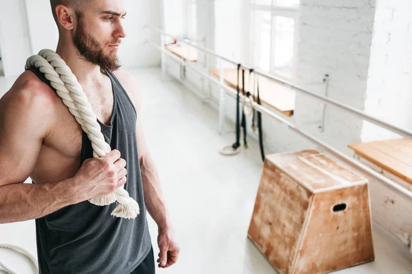 Handsome muscular man holding on shoulder rope workout in light gym. Portrait of fitness man with battle ropes at cross gym